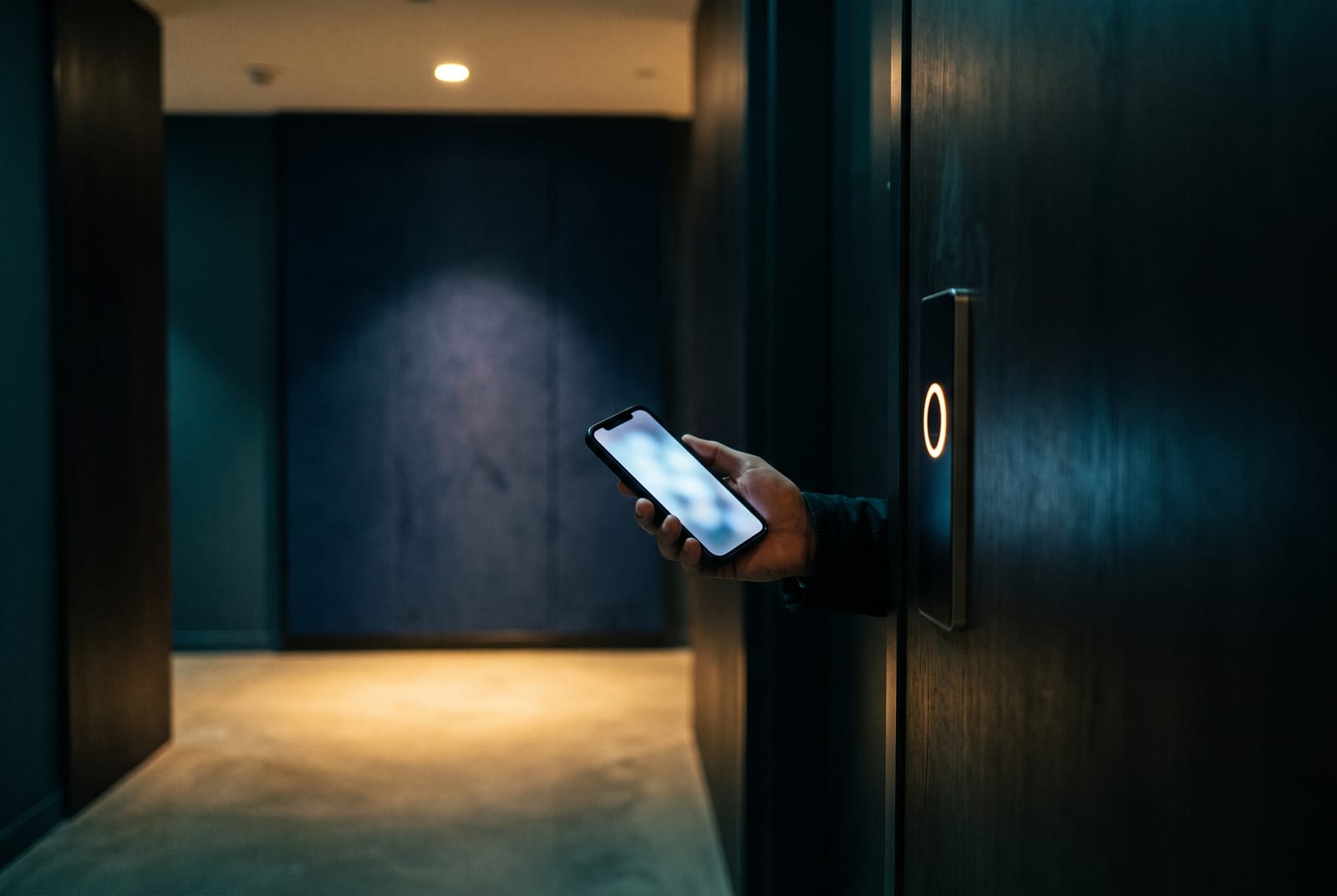A phone held up to a slim door reader in a dark modern lobby, the reader ring lit as it authorizes