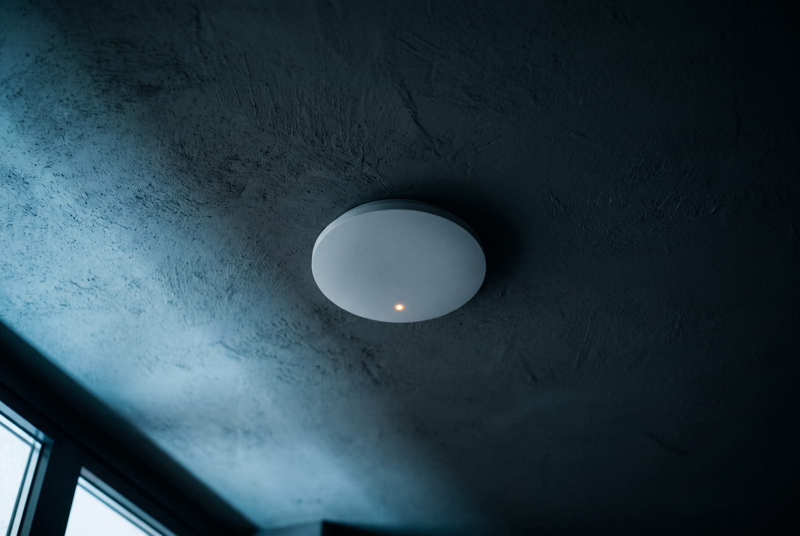 A ceiling-mounted wireless access point viewed from below, its status light glowing softly against a dark plaster ceiling